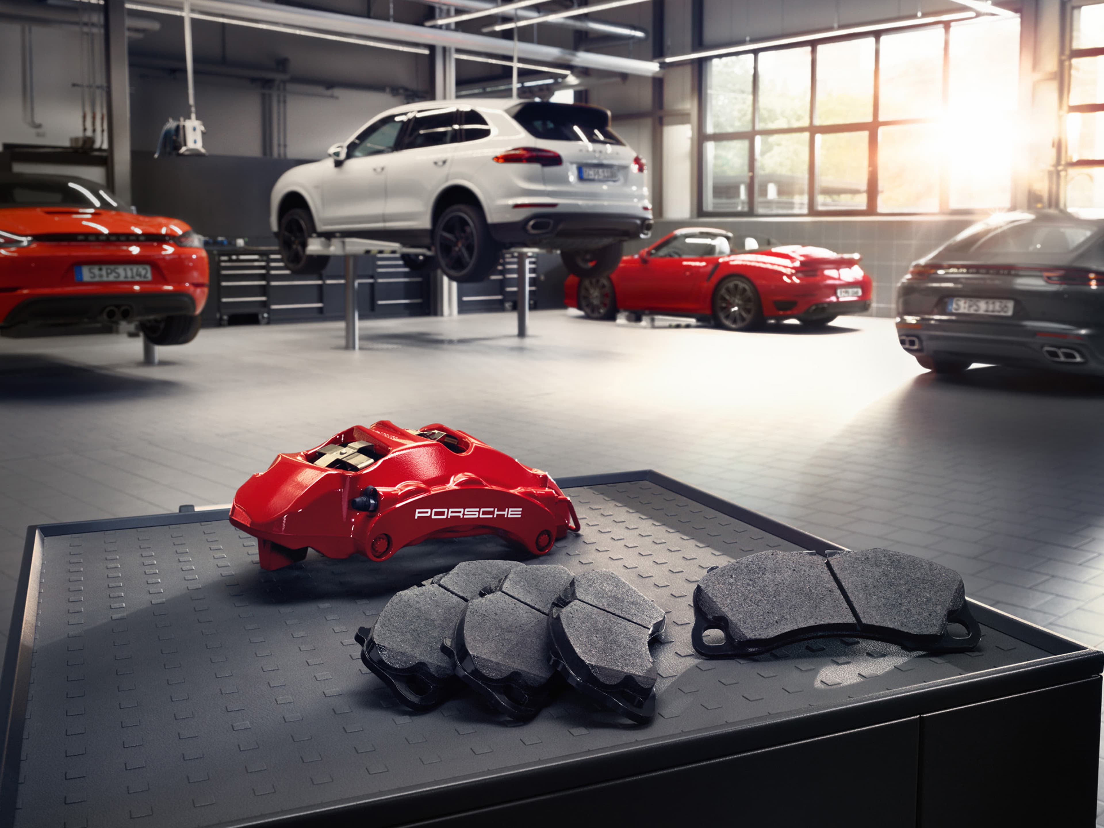 Porsche Service Center with a white Cayenne lifted on a platform. Red brake calipers and pads are displayed on a table in the foreground.